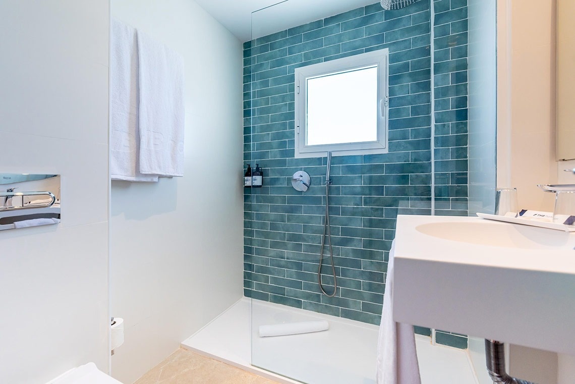 Baño moderno de hotel con ducha a ras de suelo, azulejos azules y lavabo blanco.