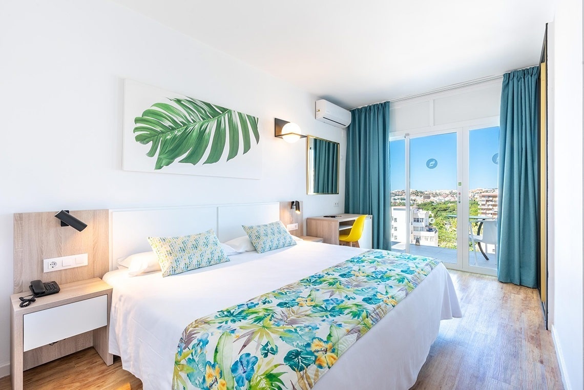 Habitación de hotel luminosa con cama doble, decoración tropical, balcón y vista a la ciudad.