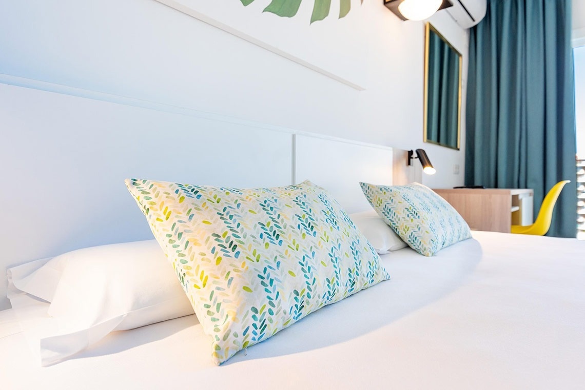 Cama de hotel con sábanas blancas, almohadas decoradas con patrón de hojas, cabecero y luz de lectura.