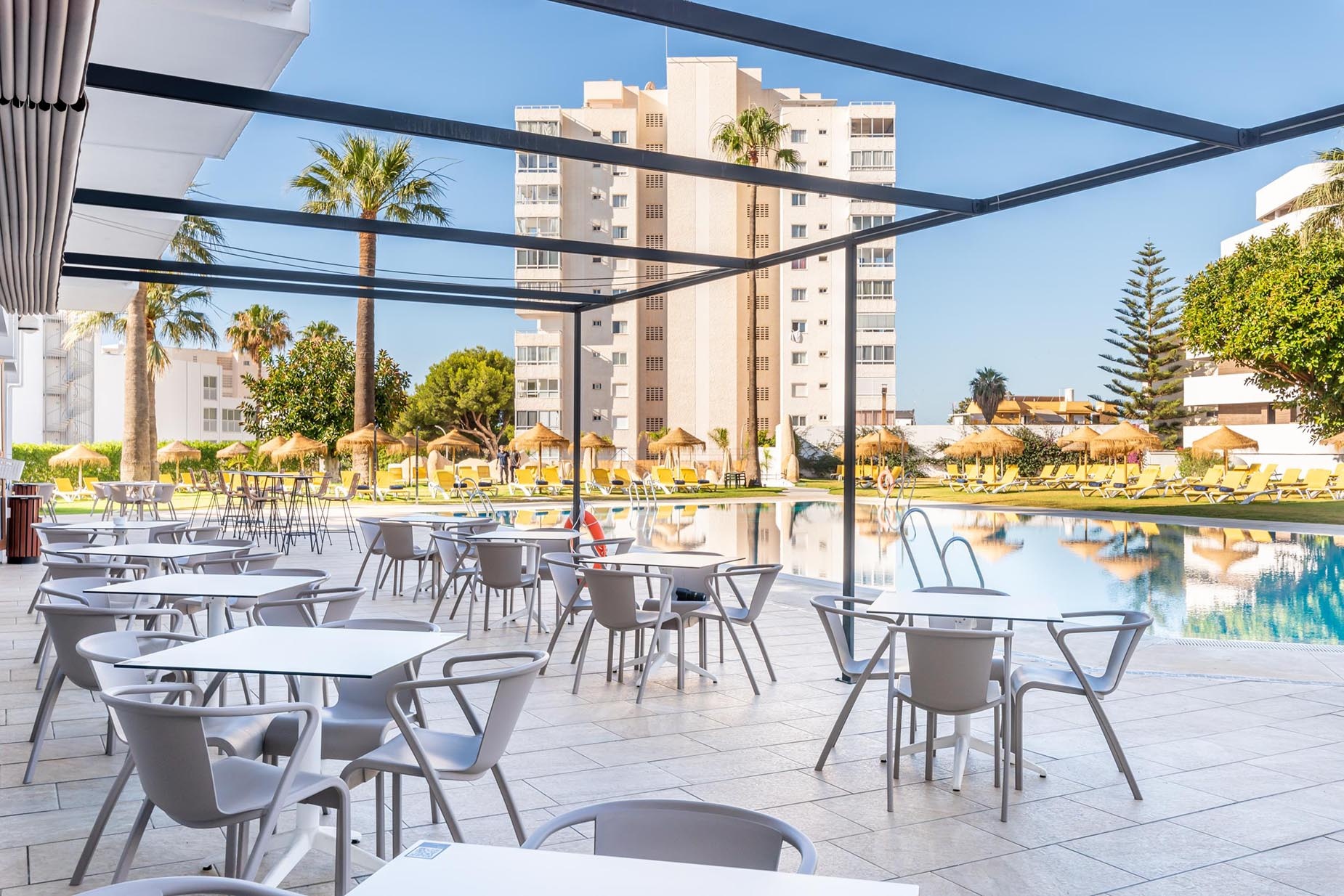 Dorobe: Hotel San Fermín. Mesa con sillas frente a piscina y edificio con palmeras.