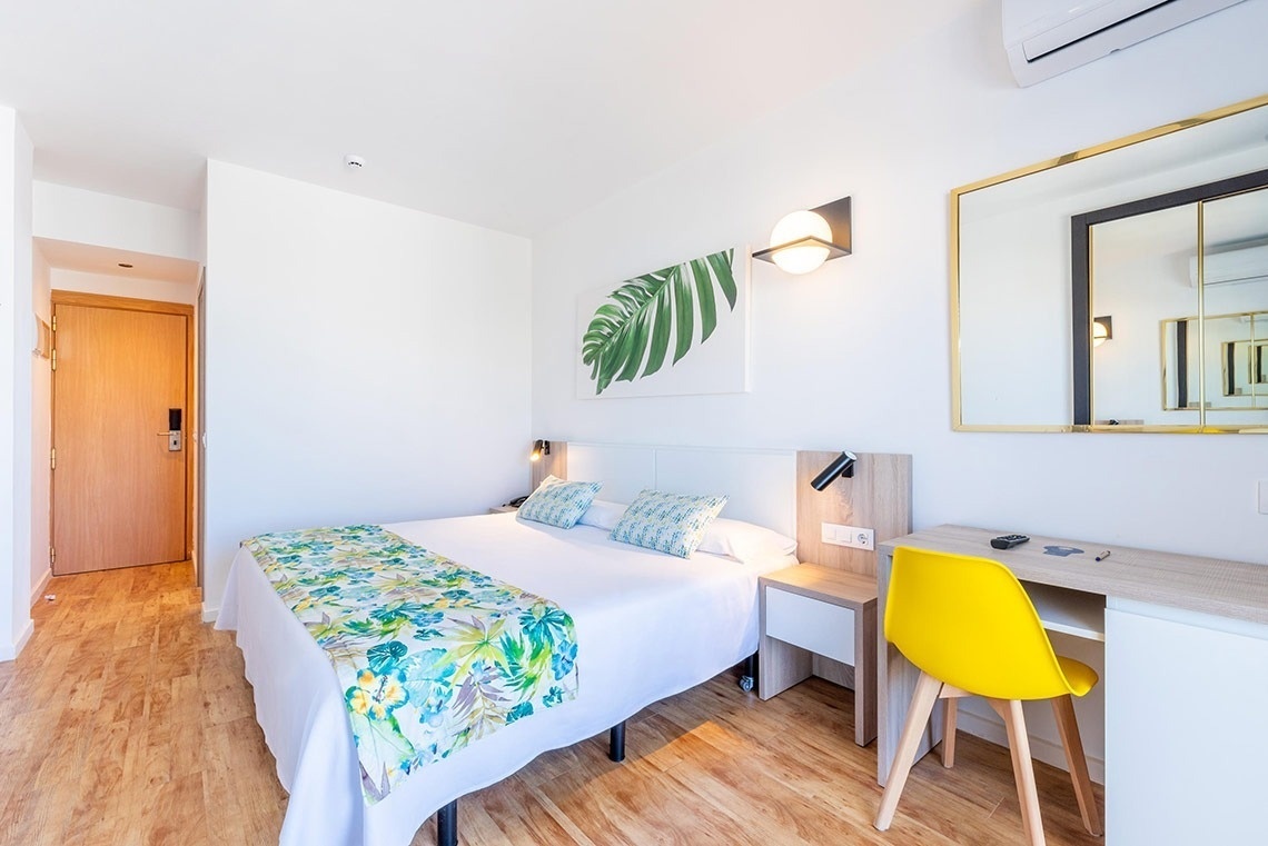 Habitación de hotel moderna con cama doble, decoración tropical, escritorio y silla amarilla.
