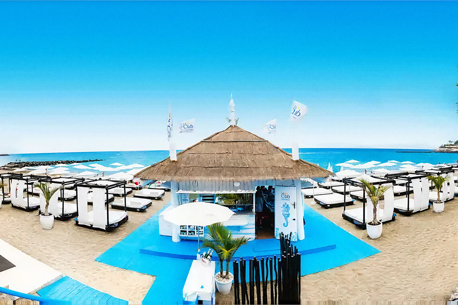 Ein Strandbild zeigt einen hellblauen Himmel über dem Meer, eine strohgedeckte Strandbar, die von einem blauen Holzsteg und zahlreichen weißen Sonnenliegen auf dem Sand umgeben ist.