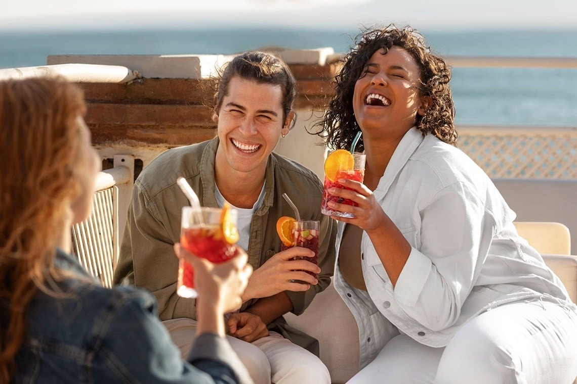 Huéspedes riendo con cócteles en terraza de hotel con vista al mar.