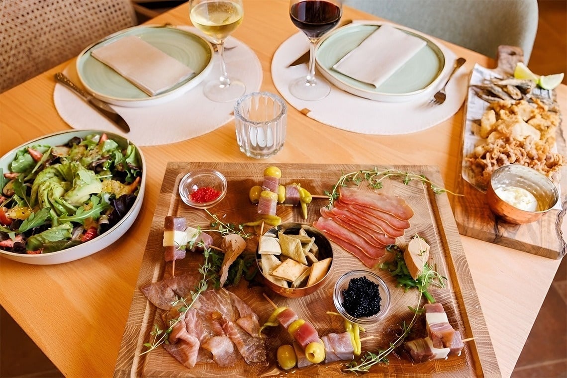 Cena gourmet en hotel con tabla de embutidos, quesos, mariscos, ensalada y vino.