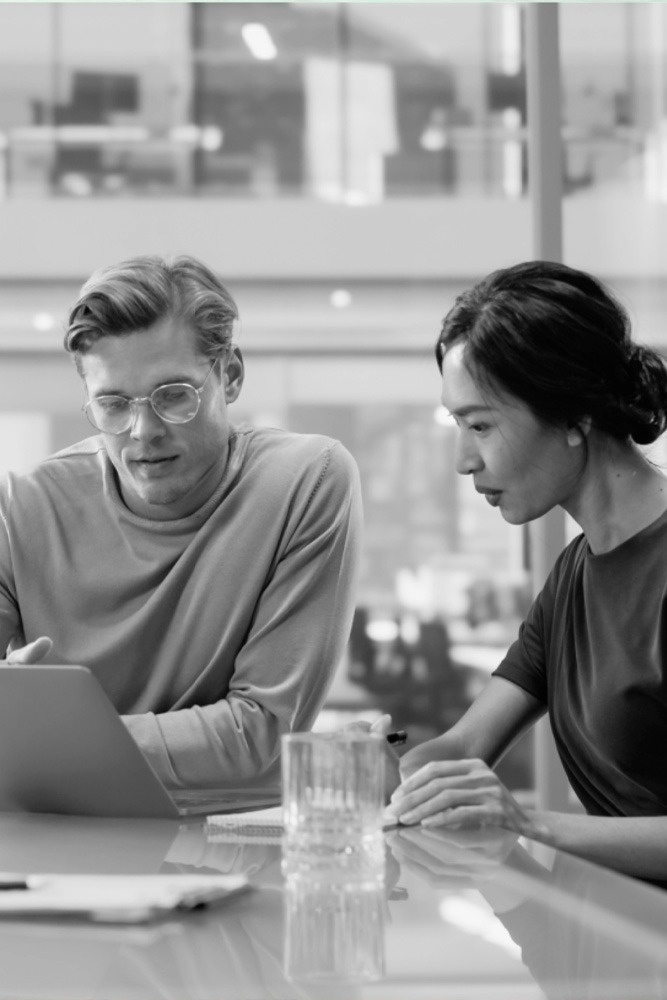 a man and a woman sit at a table looking at a laptop