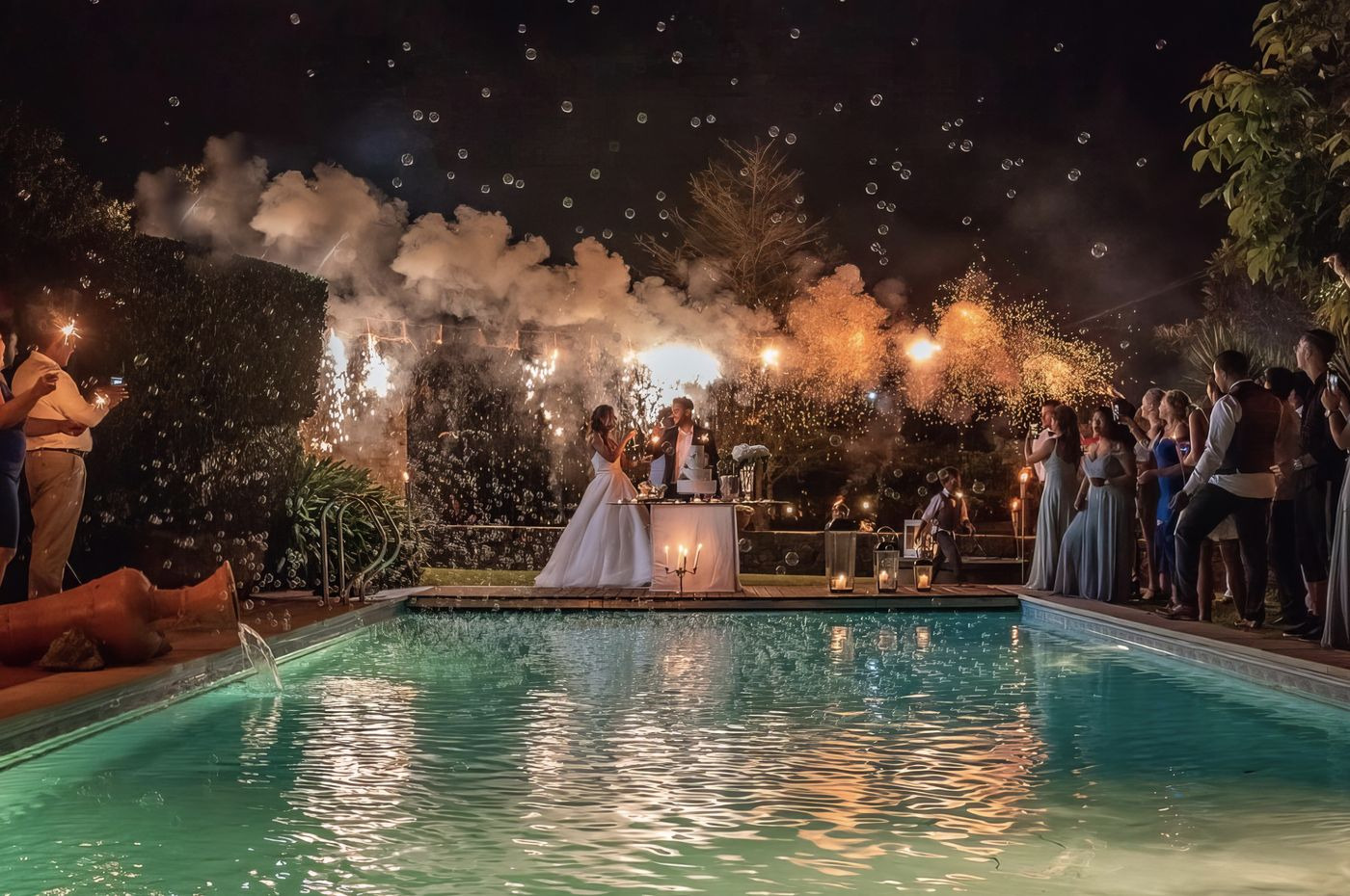 Casal junto à piscina com bolo de casamento e cascata de fadas de luz.
