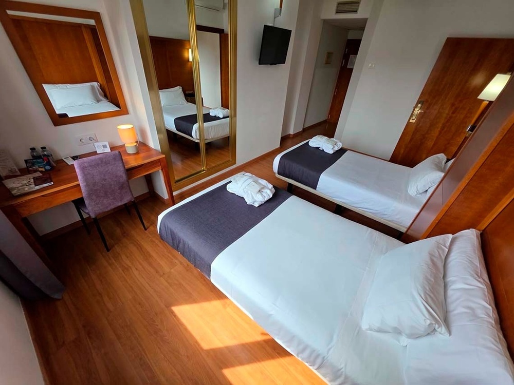 La imagen muestra una luminosa habitación de hotel con dos camas individuales, un escritorio con una silla y un gran espejo, todo sobre un suelo de madera.