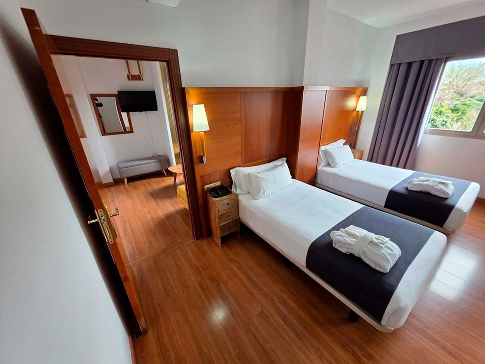 Una luminosa habitación de hotel presenta dos camas individuales con batas de baño, cabeceros de madera, y una puerta abierta hacia una zona adyacente con espejo y televisión, todo sobre un suelo de madera.