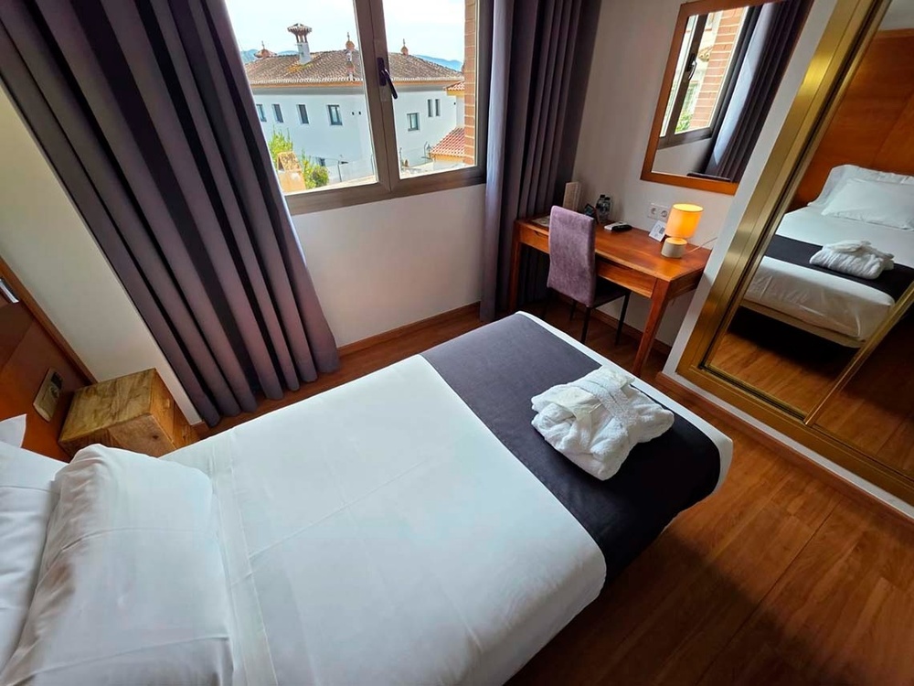 Una habitación de hotel limpia y ordenada con una cama hecha, un albornoz, un escritorio junto a una ventana con vistas a tejados y un gran espejo.