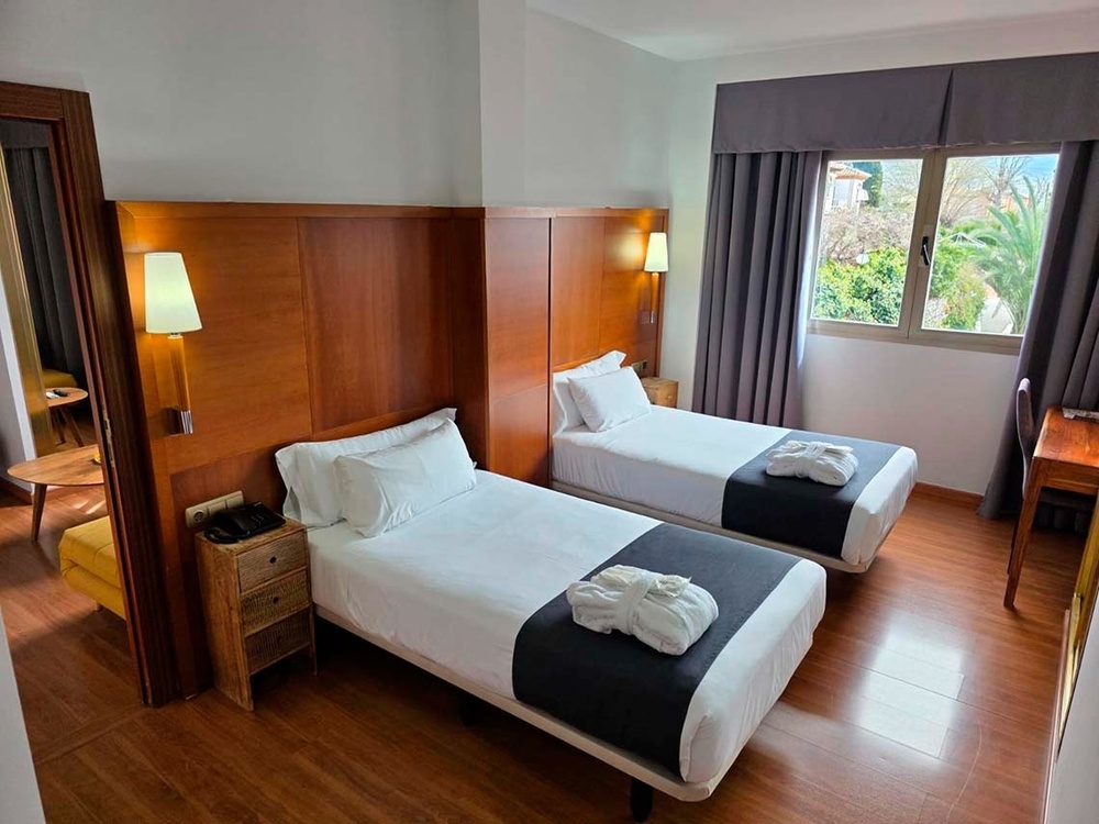 Habitación de hotel luminosa con dos camas individuales, albornoces doblados, cabecero de madera, suelo de parqué y una ventana con vistas al exterior.