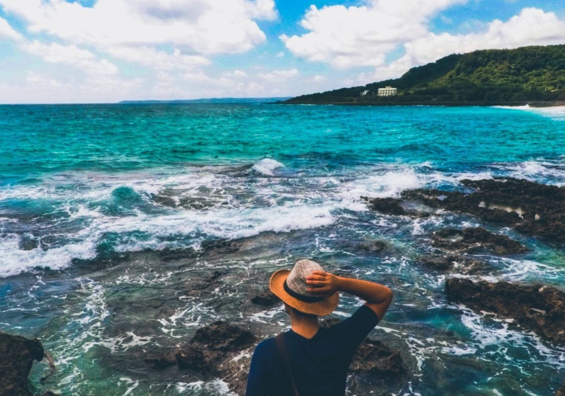 Viajar solo y elegir el destino perfecto: hombre con sombrero observa mar turquesa desde costa rocosa en Port Saplaya.