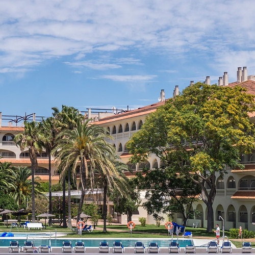 un gran edificio blanco con techo de tejas rojas está rodeado de palmeras y sillas de playa