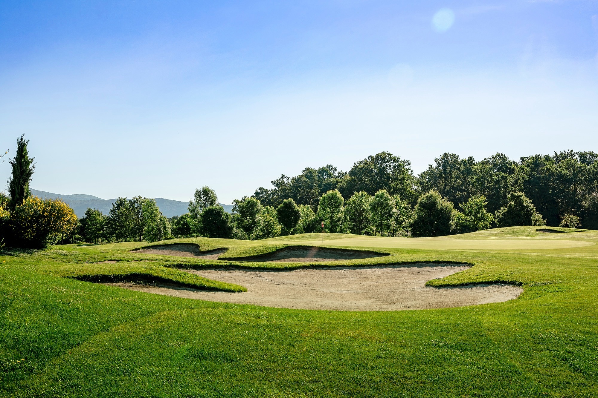 Ein Golfplatz zeigt mehrere Sandbunker und grüne Rasenflächen unter einem klaren blauen Himmel, mit Bäumen und Bergen im Hintergrund.