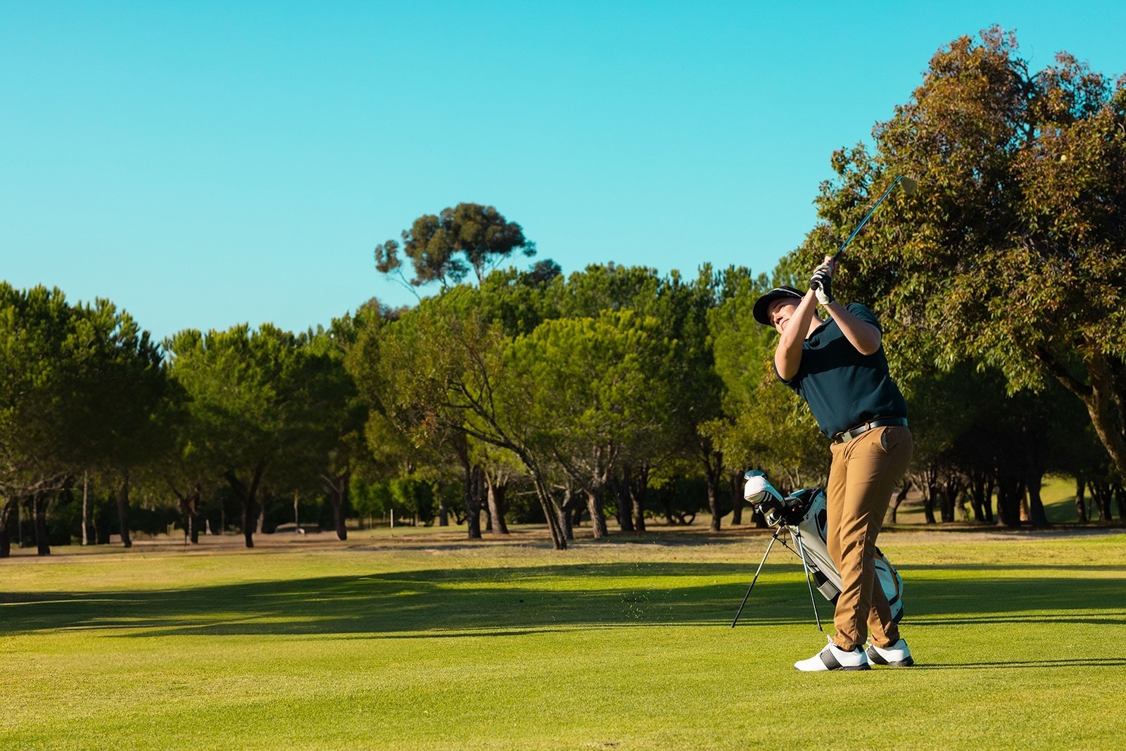 Ein Golfer schwingt seinen Schläger auf einem sonnigen Golfplatz, umgeben von grünem Rasen und Bäumen, unter einem klaren blauen Himmel.