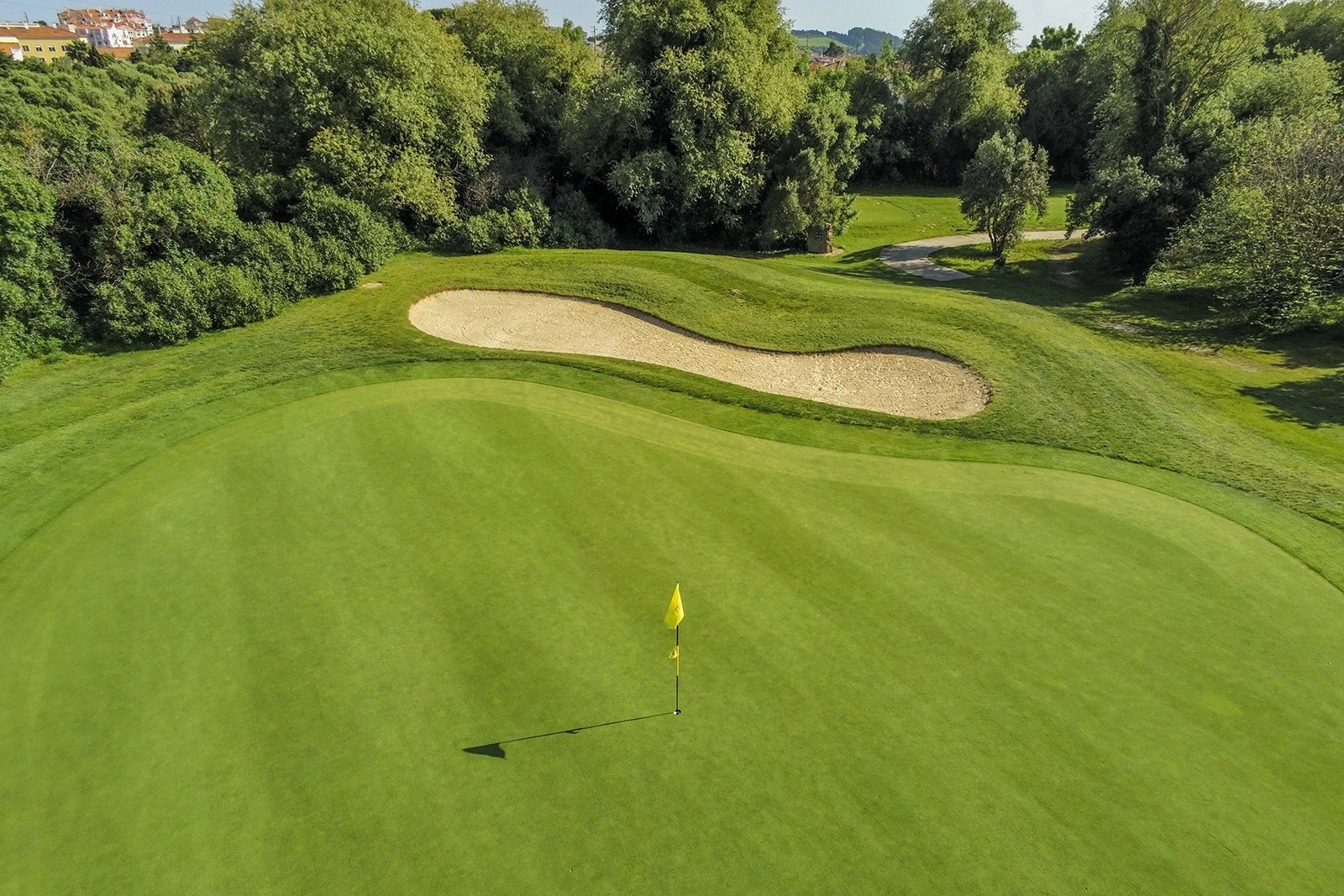 Ein grüner Golfplatz mit einer gelben Fahne auf dem Grün und einem Sandbunker im Hintergrund, umgeben von Bäumen.