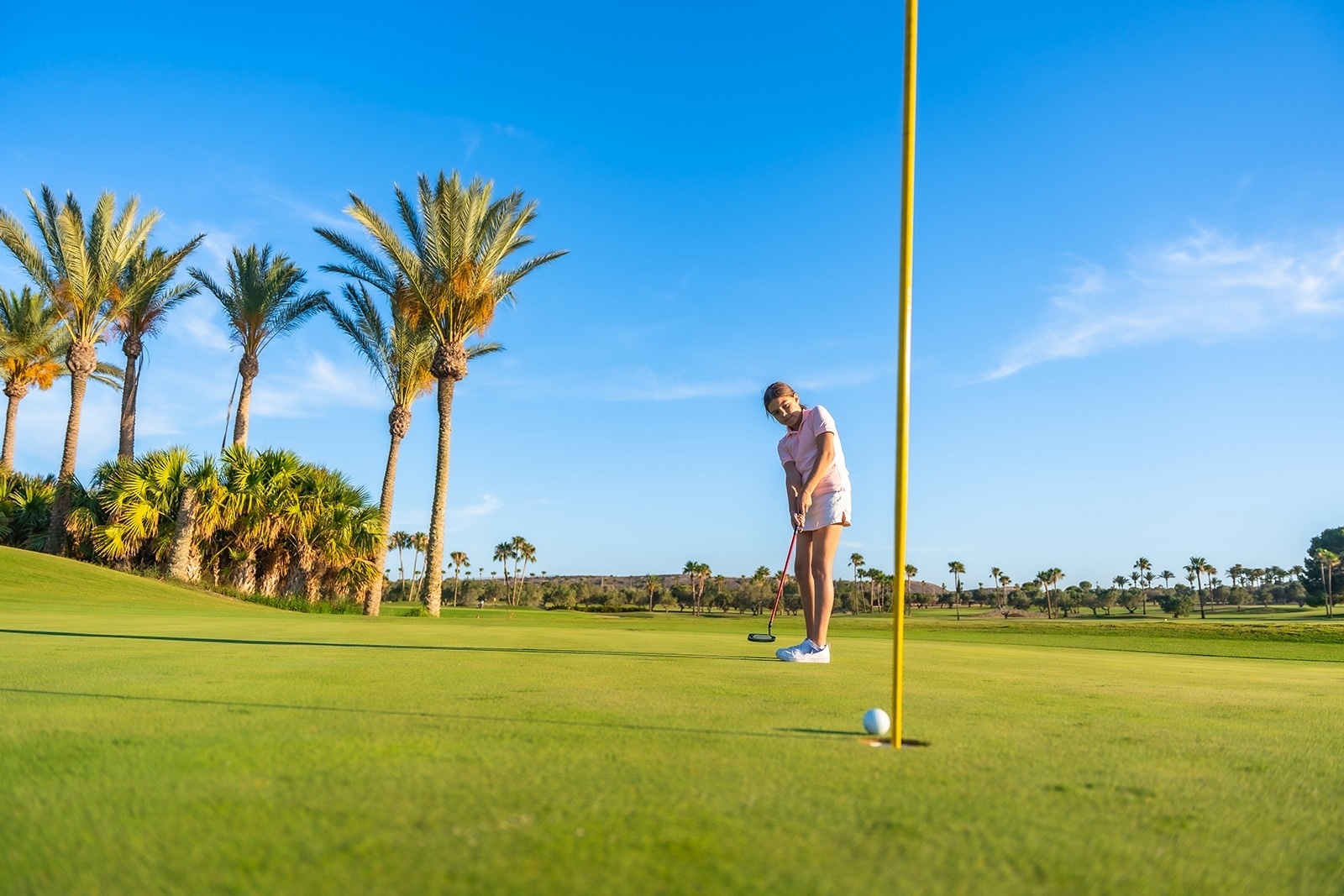 Ein junges Mädchen puttet auf einem Golfplatz mit Palmen unter blauem Himmel, wobei der Ball nahe am Loch und der gelben Flagge liegt.