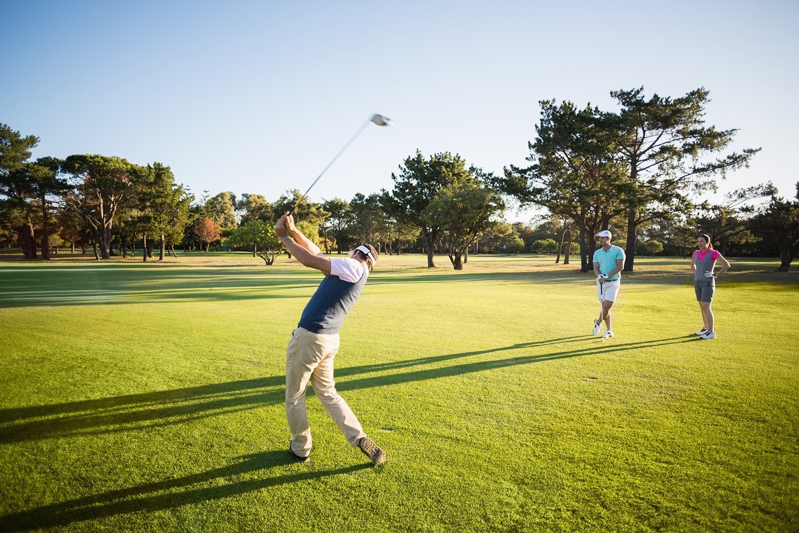 Ein Mann schlägt auf einem sonnigen Golfplatz einen Golfball, während zwei andere Golfer im Hintergrund zusehen.