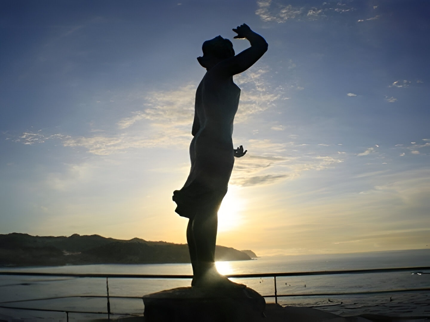 una estatua de una mujer con las manos en el aire frente al océano al atardecer