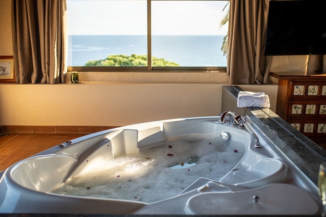 una bañera llena de espuma y flores está frente a una ventana con vista al océano