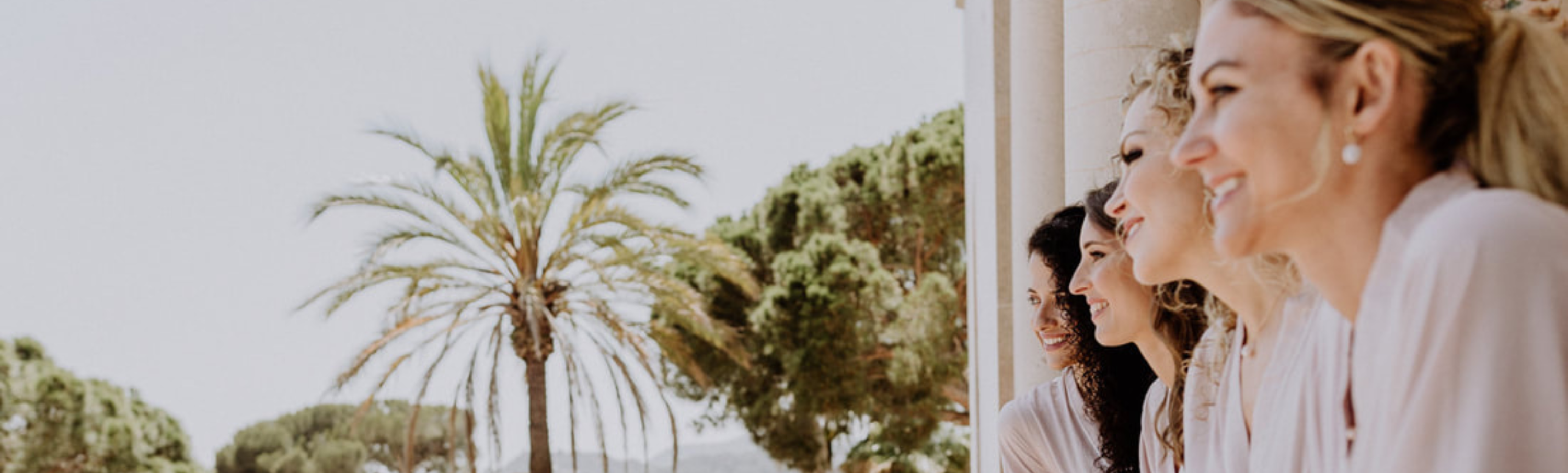 Lachende Frauen auf sonnigem Hotelbalkon mit Palmen im mediterranen Stil.