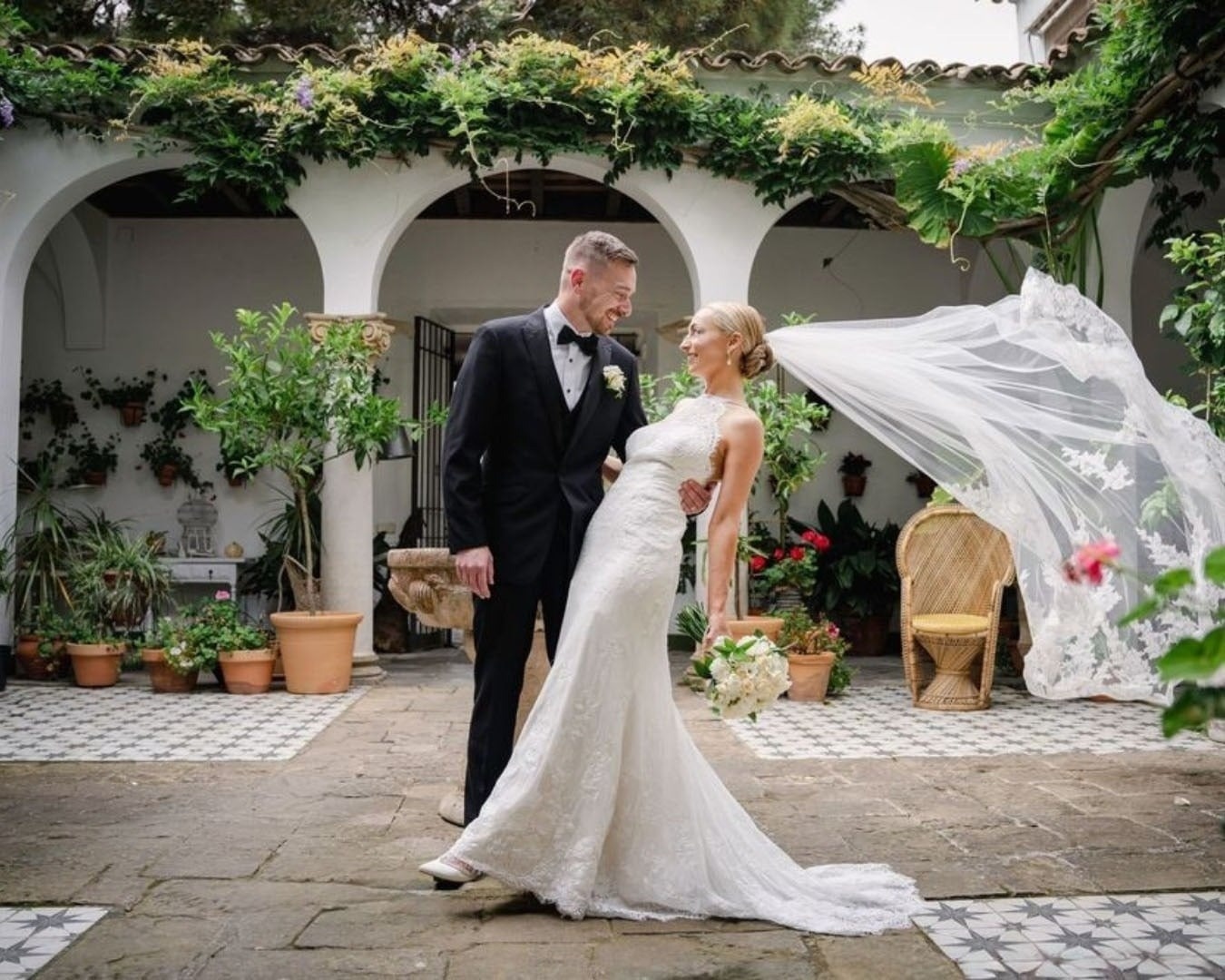 Los novios posan en un hermoso patio con arcos y vegetación, con el velo de la novia ondeando al viento.