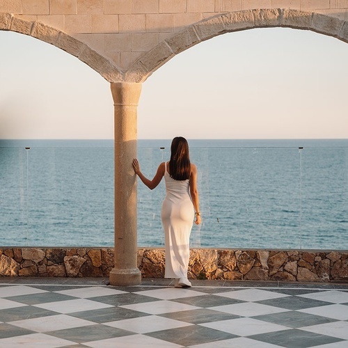 una mujer con un vestido blanco está parada en una columna mirando al océano
