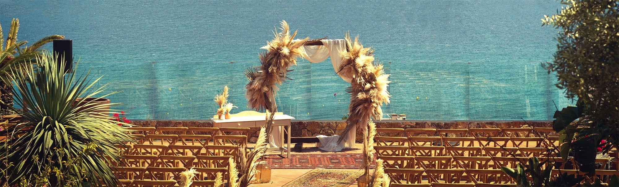Un escenari de casament exterior amb un arc de plomes de la pampa i cadires de fusta, amb el mar de fons.