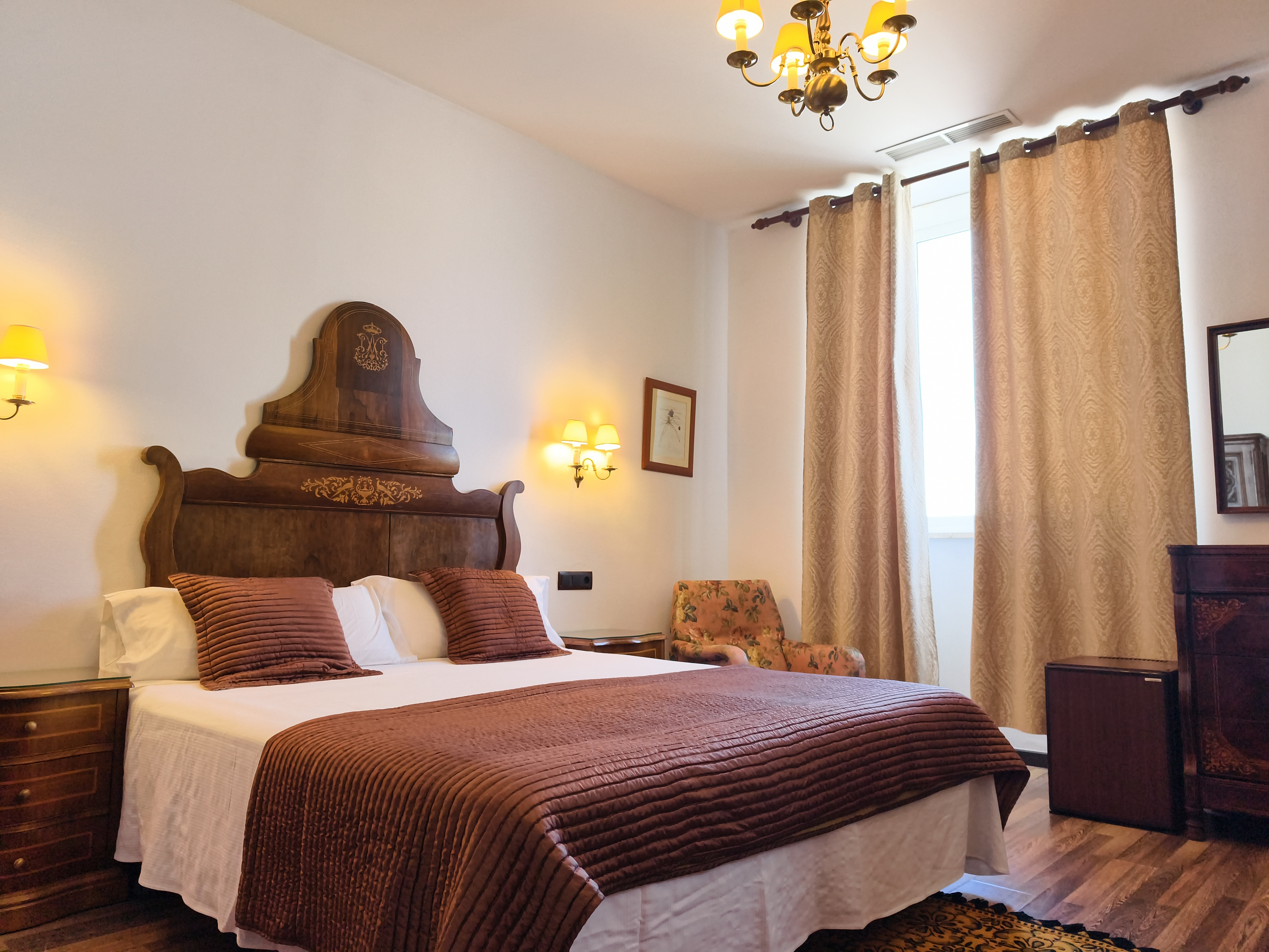 Cozy hotel bedroom with a king-size bed, dark wood headboard, plush bedding, and warm lighting. Roger De Flor By Seleqtta