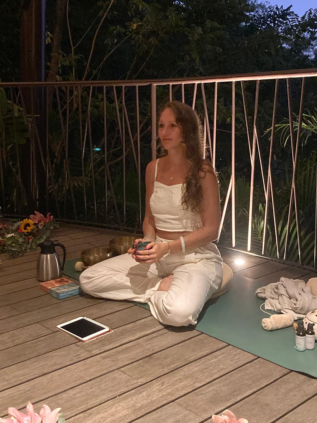 Woman meditating on a hotel balcony with sound bowls and Senhora da Rosa at dusk.