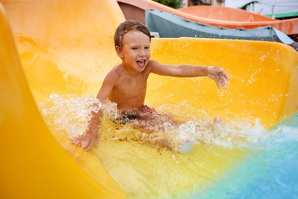 A smiling boy happily slides down a bright yellow water slide, splashing water around him.