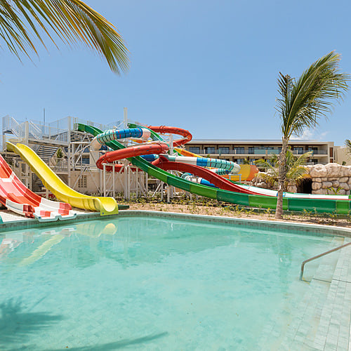 Water slides and pool at Serenade Caribe Club resort with tropical palm trees and clear blue sky.