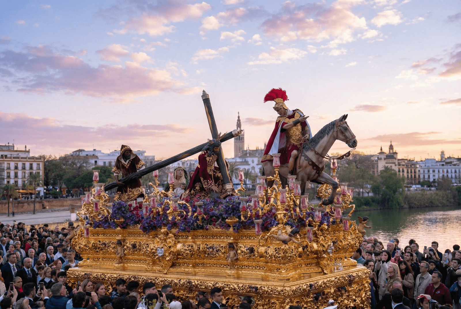 A large, ornate religious float featuring figures including Christ carrying the cross and a Roman soldier on horseback is carried through a crowded street beside a river, under a dramatic sunset sky with a city skyline in the background.