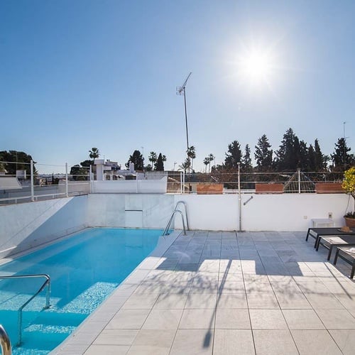 Piscina en la azotea de un hotel con zona de solárium, tumbonas y vistas al paisaje bajo un cielo soleado.