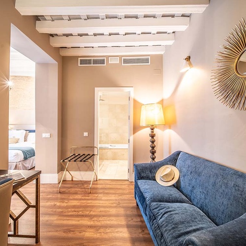Suite de hotel elegante con sofá azul, espejo sol, escritorio y entrada a dormitorio y baño.