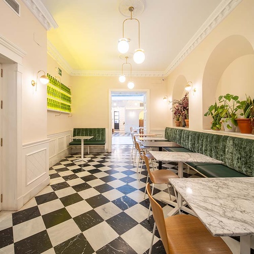 Acogedor café de hotel con banquetas verdes, mesas de mármol y suelo de cuadros. Iluminación y plantas decorativas.