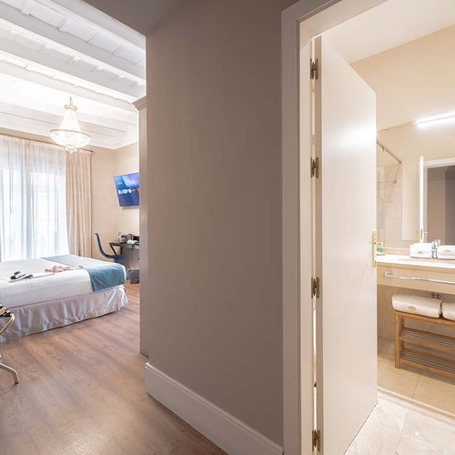 Dormitorio de hotel con cama blanca, suelo de madera y acceso a un moderno baño con tocador y ducha.