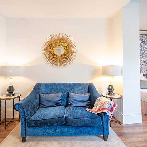 Elegante suite de hotel con sofá azul, lámparas, espejo sol y dormitorio acogedor.