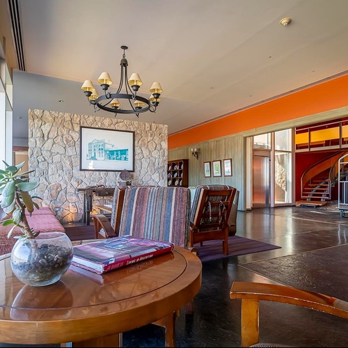 Un'ampia e luminosa area comune presenta una parete in pietra, grandi finestre, diverse poltrone e una scala a chiocciola con ascensore sullo sfondo.