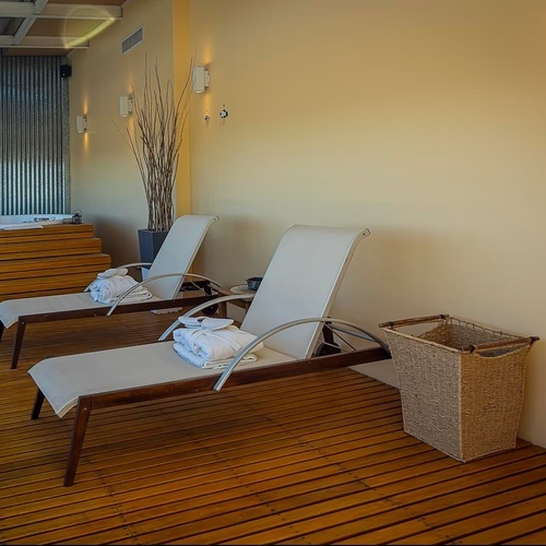 Un'area benessere interna con due lettini, accappatoi e ciabatte su un pavimento in legno, una vasca idromassaggio e una parete in metallo ondulato.