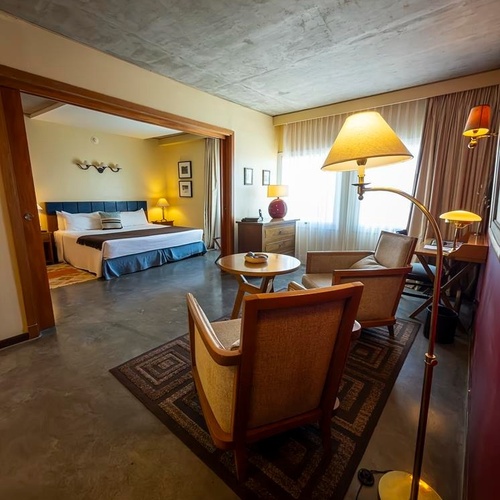 Una suite d'hotel moderna e accogliente è divisa in zona notte e area salotto, con soffitto in cemento a vista, arredi in legno e illuminazione calda.