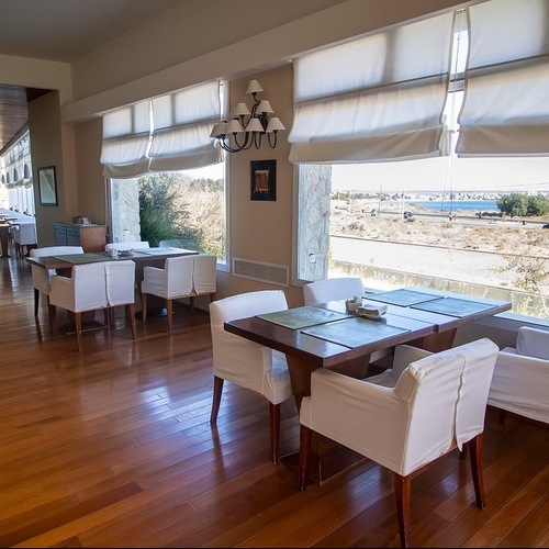 Una luminosa sala ristorante con pavimento in legno, tavoli e sedie bianche, e ampie finestre che offrono una vista su un paesaggio costiero arido con una città in lontananza.