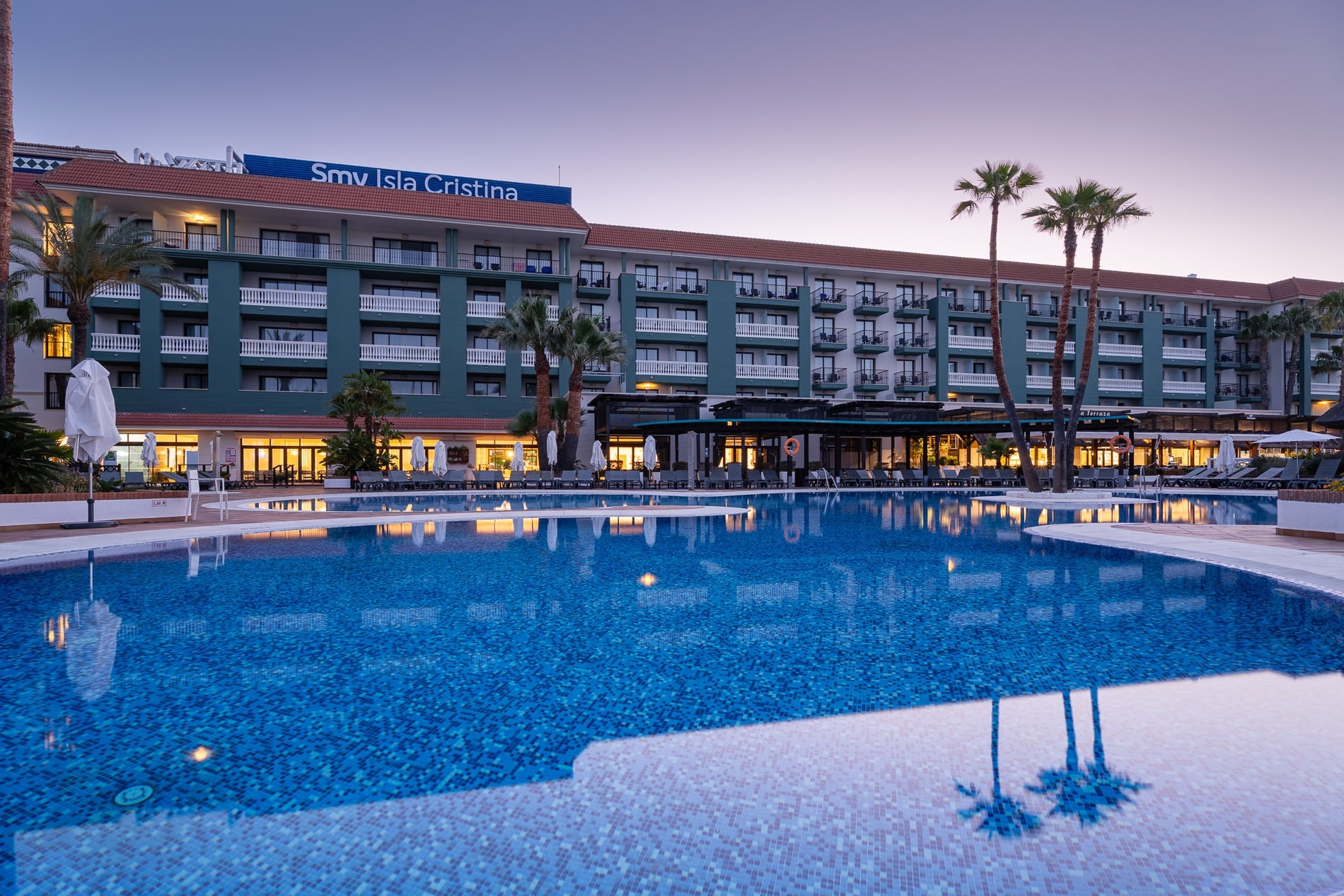 Hotel Smy Isla Cristina 4* | Official Web: Poolside view of hotel with palm trees and loungers.