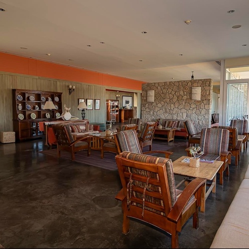 Un ampio salone in stile rustico è arredato con mobili in legno scuro, divani e poltrone con cuscini a fantasia, pareti rivestite in pietra e legno, e un lungo divano chiaro accanto a grandi finestre.