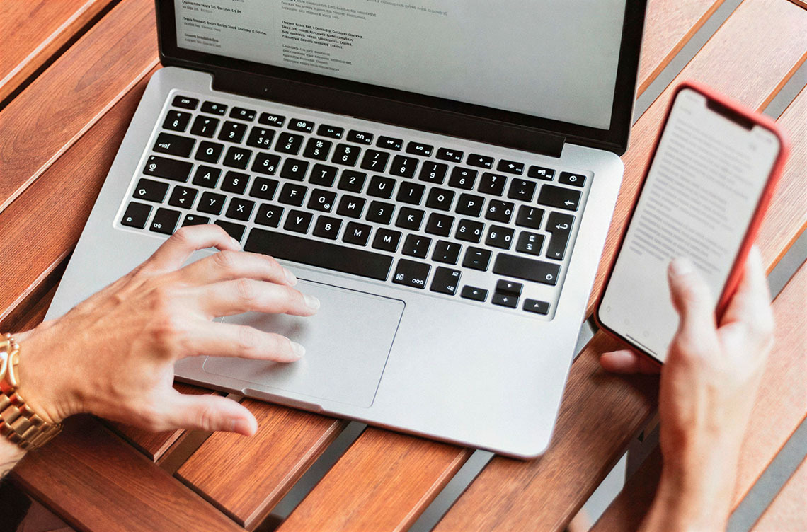 Person using laptop and phone on a wooden table, ideal for Smy: Corporativa website.