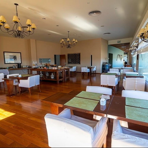 Una spaziosa sala da pranzo o ristorante con pavimenti in legno lucidi, tavoli apparecchiati con tovagliette verdi, sedie con coperture bianche, un'area buffet e lampadari, illuminata da finestre che danno sull'esterno.