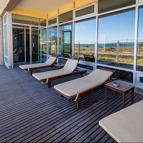 L'immagine mostra una spaziosa terrazza con pavimento in legno e lettini prendisole, con ampie finestre che riflettono il cielo e un panorama marittimo.