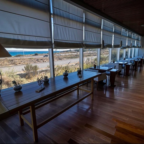 Un moderno ristorante con ampie vetrate si affaccia sul mare e su una strada costiera, caratterizzato da un arredamento in legno e tessuti neutri.