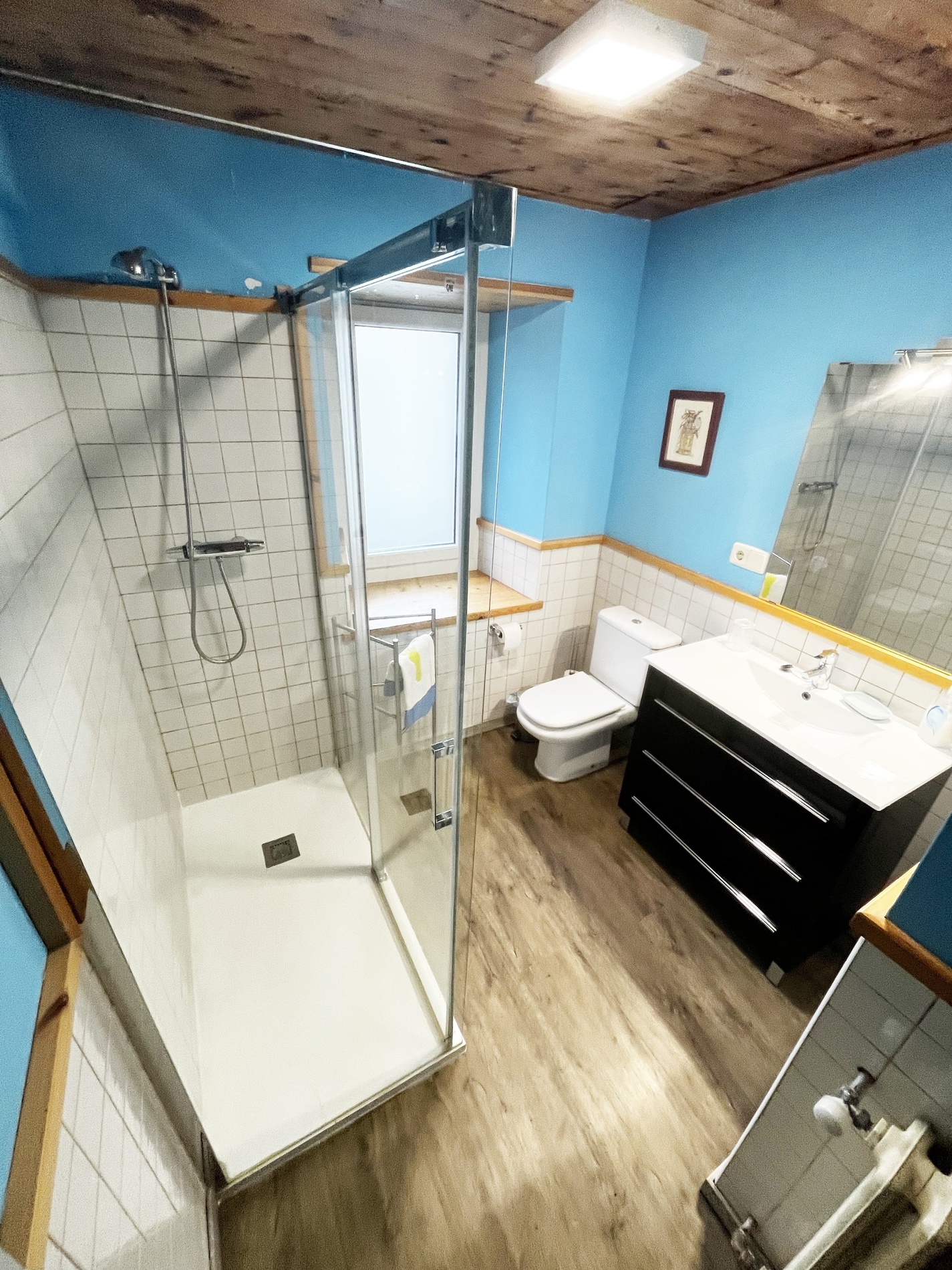 Cette image présente une salle de bain moderne avec des murs bleus et carrelés blancs, un plafond et un sol en bois, une douche vitrée, un lavabo noir et des toilettes.