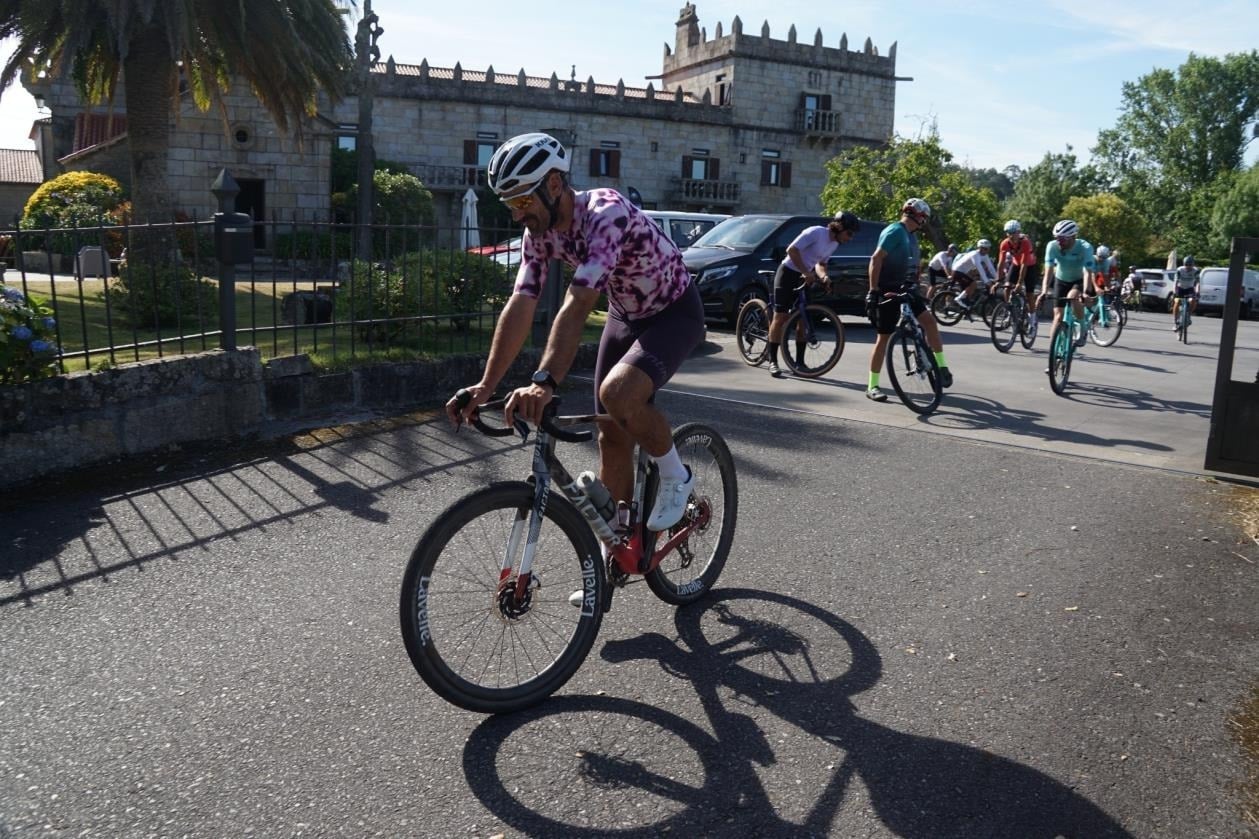 Cyclists ride past a historic stone building, possibly a SNO: Corporativa.