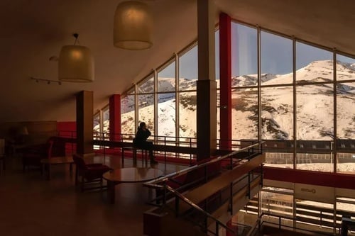 a woman sits in front of a large window looking out to a snowy mountain